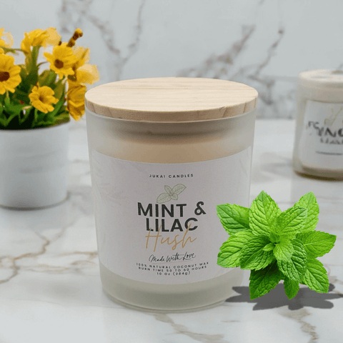 Candle labeled 'Mint & Lilac' with a wooden lid on a marble surface, surrounded by yellow flowers and mint leaves.